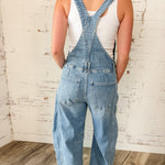 Person wearing denim overalls with a white tank top against a rustic brick wall.