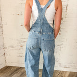 Person wearing denim overalls with a white tank top against a rustic brick wall.