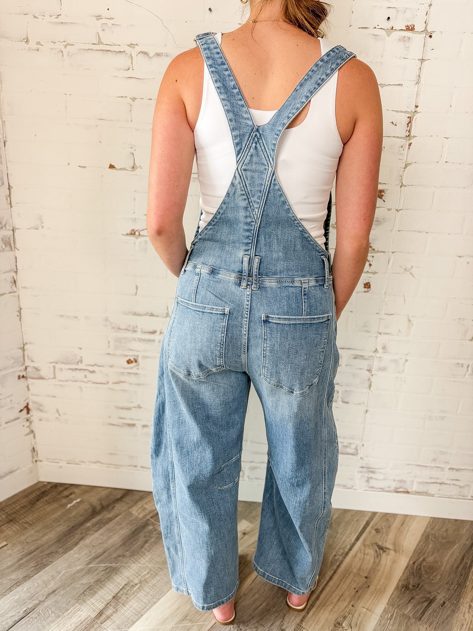 Person wearing denim overalls with a white tank top against a rustic brick wall.